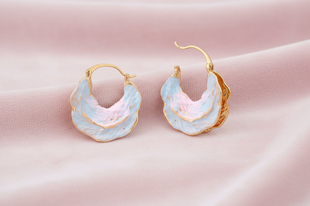 Shell-shaped hoop earrings with iridescent blue-pink enamel and gold trim on cream background