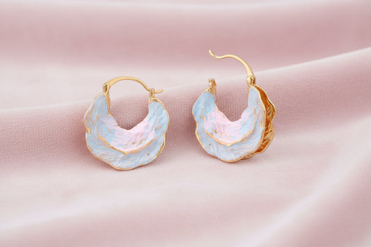 Shell-shaped hoop earrings with iridescent blue-pink enamel and gold trim on cream background