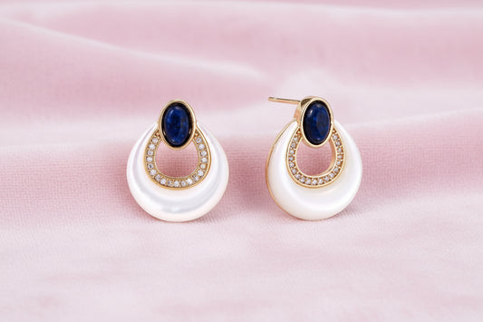 Gold drop earrings with oval lapis lazuli, crystal-paved frame, and iridescent mother-of-pearl teardrop on cream background