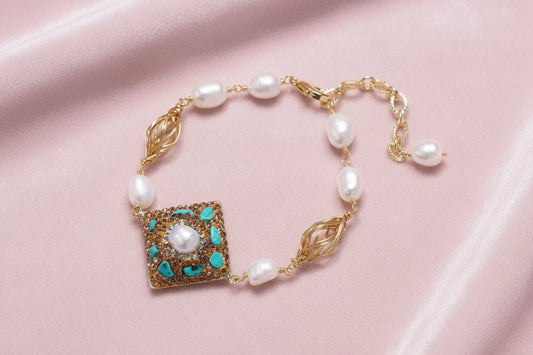 Gold-plated bracelet with baroque centerpiece, turquoise stones, freshwater pearls, and ornate chain links on cream background.