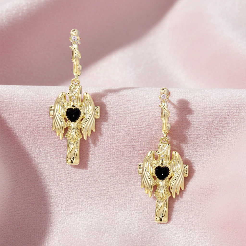 Gold angel wing drop earrings with black heart-shaped zirconia center and twisted posts on beige background