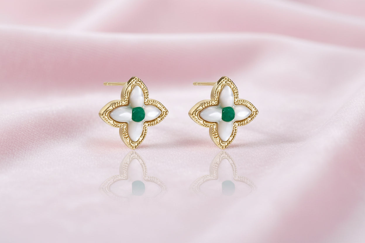 Gold quatrefoil stud earrings with white mother-of-pearl petals and emerald green gemstone center on beige background.