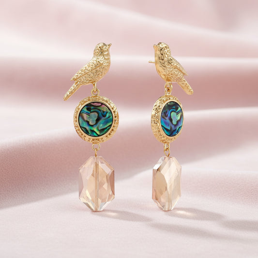 Gold-plated bird stud earrings with iridescent abalone shell medallions and champagne crystal drops on cream background