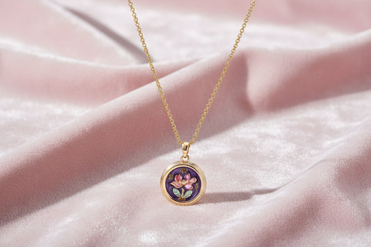Gold-plated circular pendant with hand-painted purple enamel floral design on delicate chain against cream background