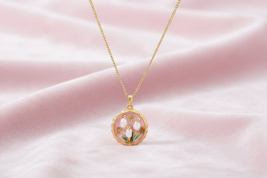 Gold pendant with pink enamel backdrop, white freshwater pearls as tulip blooms, and green leaf accents on rope chain.