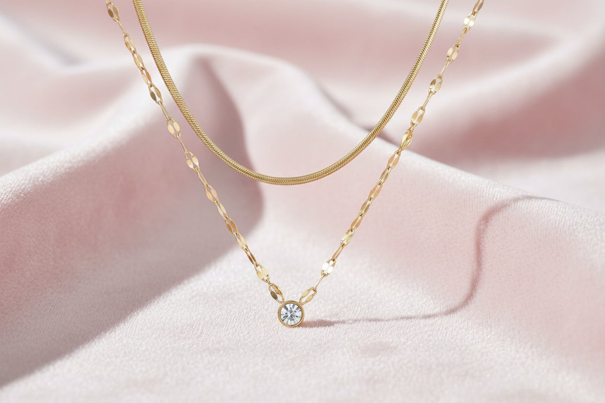 Layered gold necklace with herringbone and link chains, small bezel-set crystal pendant on cream background