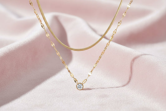 Layered gold necklace with herringbone and link chains, small bezel-set crystal pendant on cream background