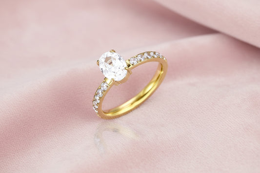 Gold-plated solitaire ring with oval zirconia center stone and pavé band on cream background