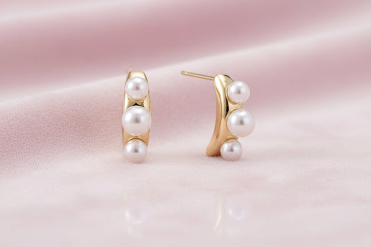 14K gold-plated curved stud earrings with three white freshwater pearls aligned vertically on cream background