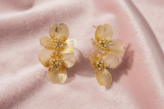 Pair of gold-plated double flower earrings with textured petals and crystal centers on cream background.