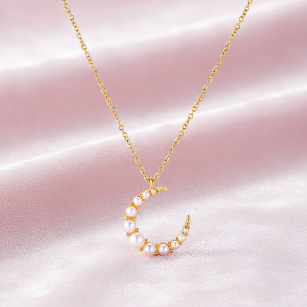 Gold crescent moon pendant with graduated white pearls on delicate chain against cream background