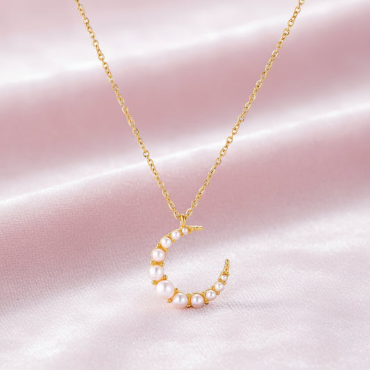 Gold crescent moon pendant with graduated white pearls on delicate chain against cream background