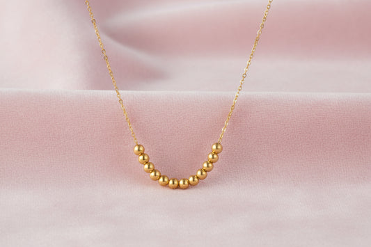 Delicate gold-plated necklace with graduated spherical beads forming gentle smile curve on beige background
