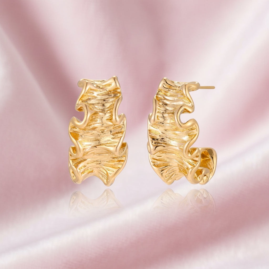 Pair of gold huggie hoops with sculpted wave texture and dimensional ripples on neutral background.