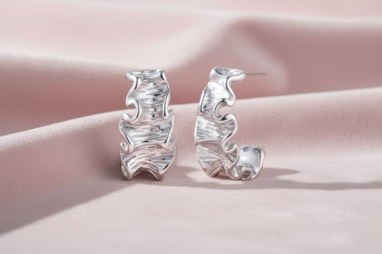 White gold plated sculptural hoop earrings with organic wavy edges and reflective metallic finish on cream background