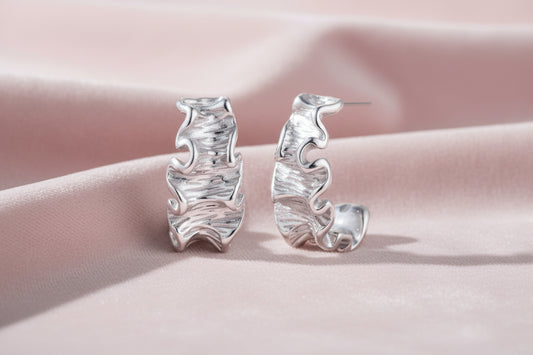 White gold plated sculptural hoop earrings with organic wavy edges and reflective metallic finish on cream background