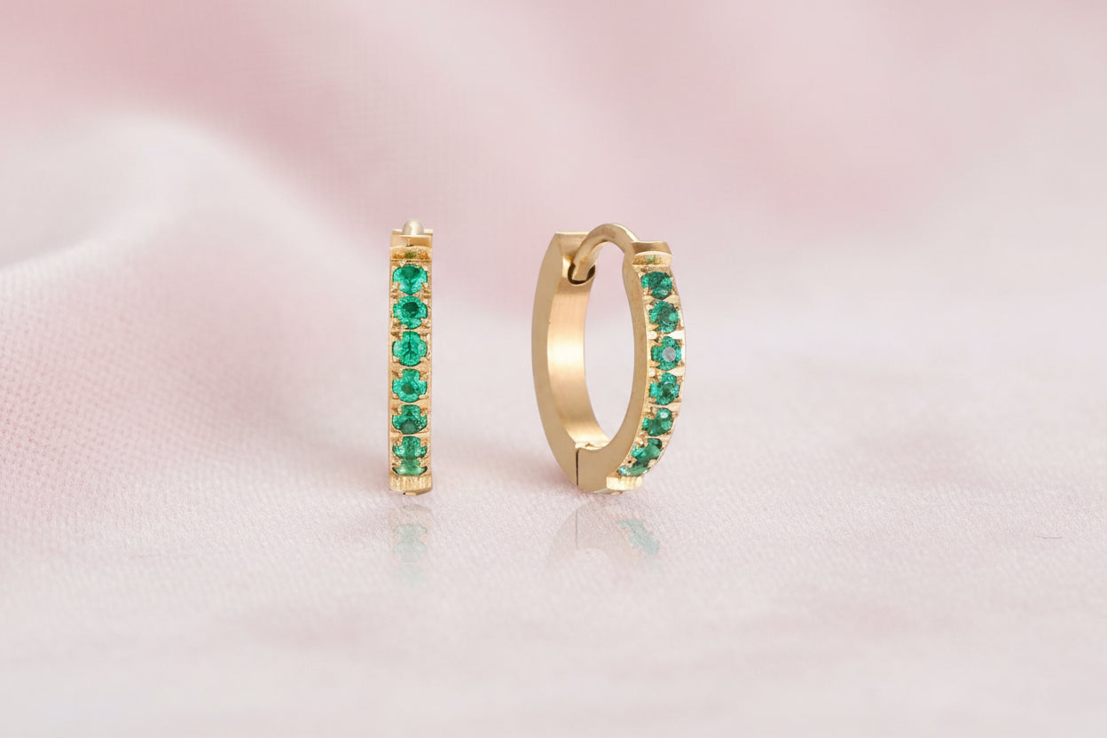 Pair of small gold huggie hoop earrings with bright green cubic zirconia stones on cream background