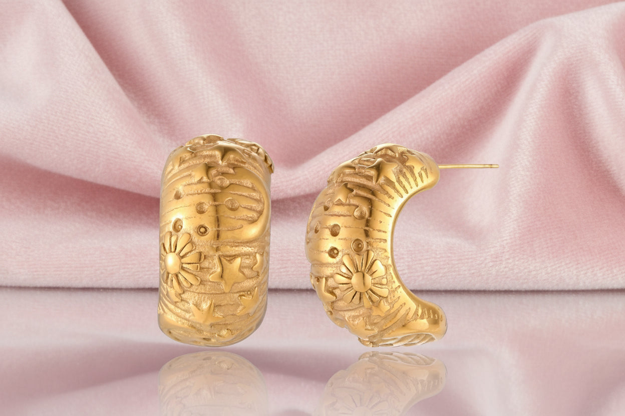 Wide gold hoop earrings with embossed stars and sun motifs on cream background, showing dimensional relief details.