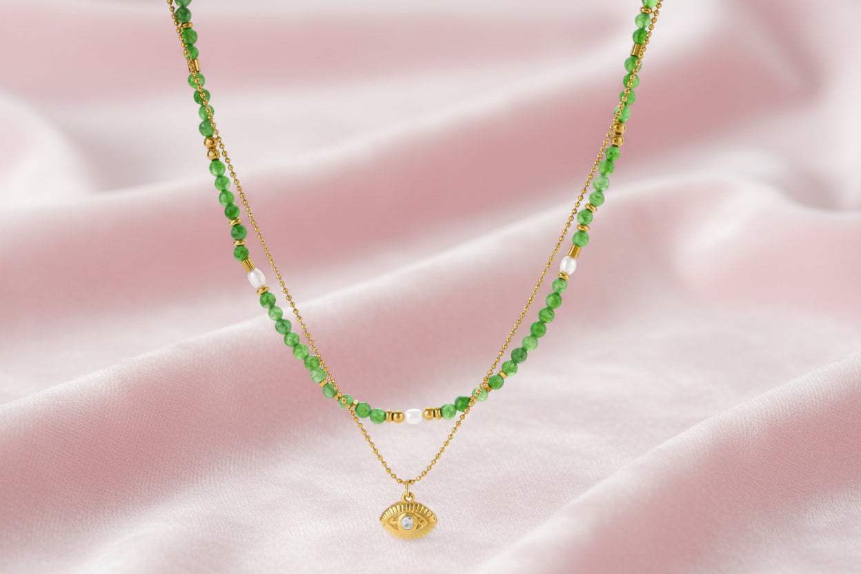 Layered necklace with green stone beads, gold spacers, white accents, and protective eye pendant on cream background.