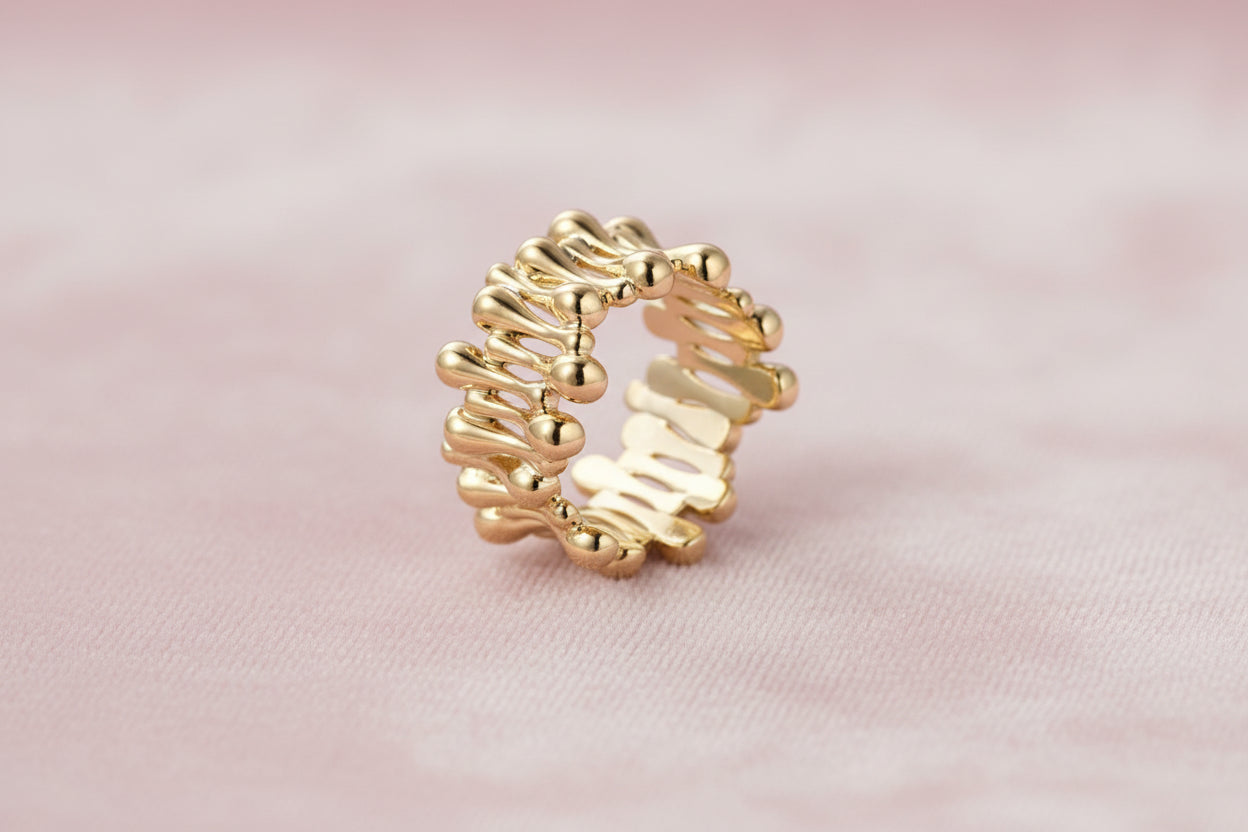 Gold-plated brass ring with sculptural petal-like elements in a wide band design on beige background