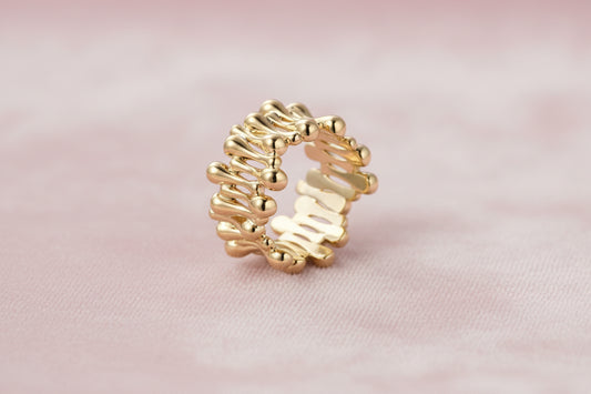 Gold-plated brass ring with sculptural petal-like elements in a wide band design on beige background