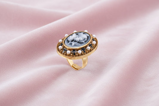 Gold-plated ring with oval blue floral cameo, pearl accents, and black enamel detailing on white background.